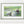 The photo of a frog and becorn looking at each other in a thin, sage green frame on a white shelf against a white wall. On either side of it are potted plants and a small stack of books. In the lower right corner of the print’s white border is David Bird’s signature.