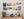 Eight metal prints mounted without frames in two rows on a pale gray wall. In the first row are “Glowdrop”, “Welcoming Party?”, “Forsythia”, and “Cardinal”. In the second row are “The Offering”, “Winter Female Cardinal”, “Frog”, and “Tulip”. In the lower right corner of the prints, David Bird’s signature is visible. Underneath them is a sideboard with several potted plants.